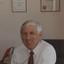 John Cook sitting in his office chair with framed certificates behind him
