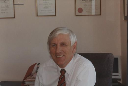 John Cook sitting in his office chair with framed certificates behind him