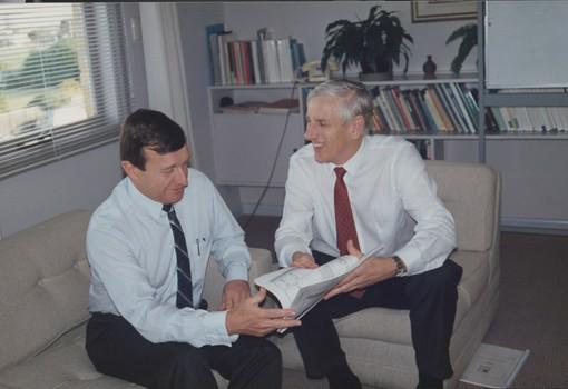 Two men sit on lounge chairs looking at paperwork and smiling 