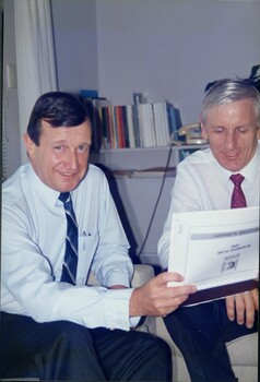 Two men sit on lounge chairs looking at paperwork and smiling 