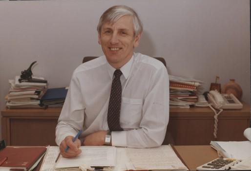 Smiling to camera as he sits at his desk surrounded by paperwork