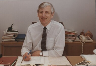 Smiling to camera as he sits at his desk surrounded by paperwork
