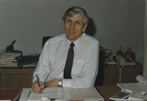 Looks to camera as he holds a pen above a letter surrounded by paperwork