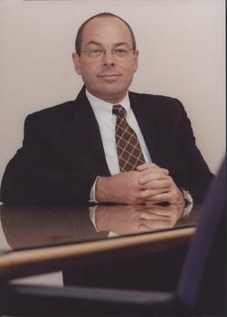 Suited man sits with hands held resting on glass topped desk
