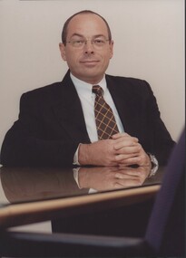 Suited man sits with hands held resting on glass topped desk