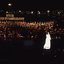 Stella Axarlis in white dress sings to crowd whilst behind her the choir and band perform