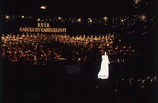 Stella Axarlis in white dress sings to crowd whilst behind her the choir and band perform