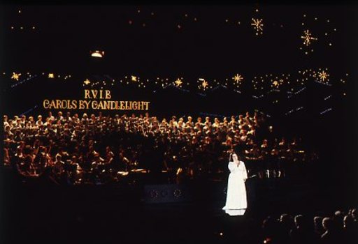 Stella Axarlis in white dress sings to crowd whilst behind her the choir and band perform
