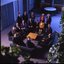 15 people sit around a small table and turn towards the photographer who is a floor above them