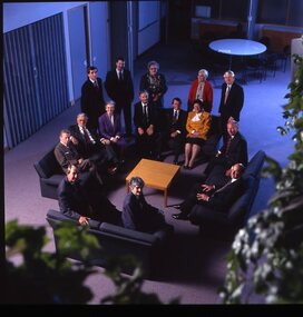 15 people sit around a small table and turn towards the photographer who is a floor above them