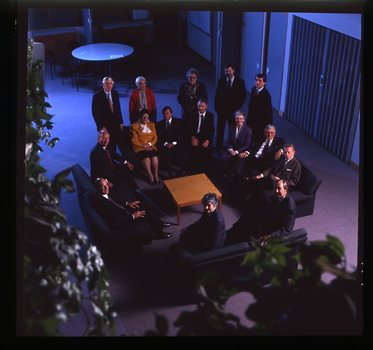 15 people sit around a small table and turn towards the photographer who is a floor above them