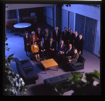 15 people sit around a small table and turn towards the photographer who is a floor above them