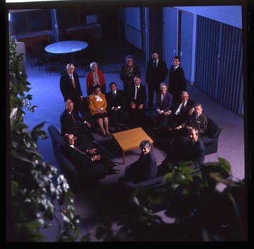 15 people sit around a small table and turn towards the photographer who is a floor above them