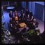 15 people sit around a small table and turn towards the photographer who is a floor above them