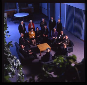 15 people sit around a small table and turn towards the photographer who is a floor above them