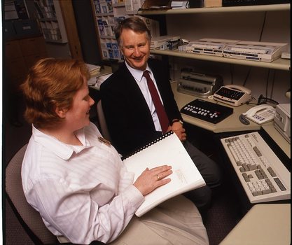 Robin Pleydell sits next to AFB Staff member at her workstation whilst she is reading Braille