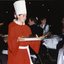 A chorister wearing a chef's hat holds a plate with a Christmas pudding