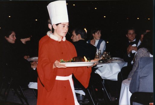 A chorister wearing a chef's hat holds a plate with a Christmas pudding
