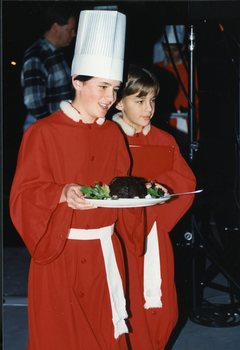 A chorister wearing a chef's hat holds a plate with a Christmas pudding accompanied by another chorister