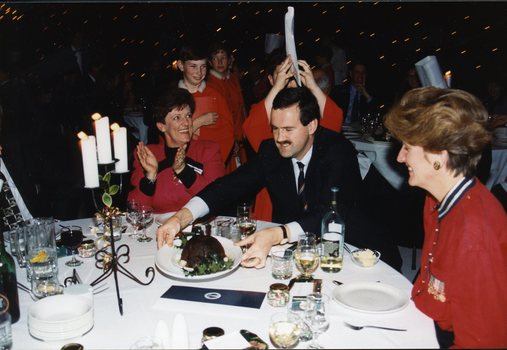Carol O'Reilly claps as man puts Christmas pudding on table and chorister is about to place chef's hat on man's head