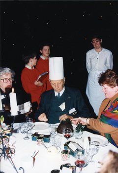 Man in centre wears a paper chef's hat, whilst the woman helps cut up the Christmas pudding