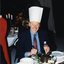 Man wearing a tall paper chef's hat, smiles for the camera as he cuts the Christmas pudding