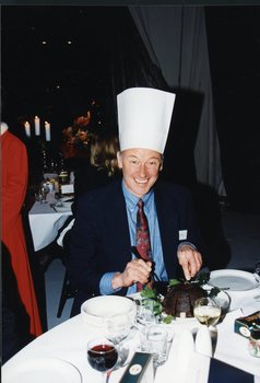 Man wearing a tall paper chef's hat, smiles for the camera as he cuts the Christmas pudding