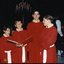 Four Choristers stand, one holding a song book