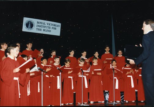 National Boys Choir dressed in red, with their conductor, in front of a starry backdrop and RVIB sign