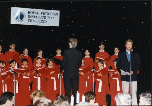 National Boys Choir dressed in red, with their conductor, in front of a starry backdrop with Peter Cupples