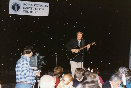 Peter Cupples on stage with guitar and a cameraman recording