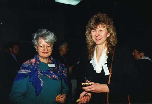 Two females stand and turn towards camera with a drink or an appetizer in their hand