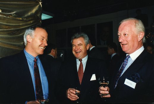 Three men stand and smile as they hold glasses of wine