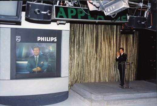 Pete Smith claps as Ray Martin appears on a giant television next to him.