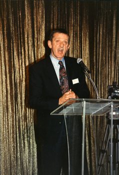 Acting CEO Len Evans gives a welcome address in front of a gold curtain