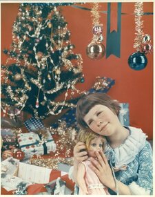 Girl in blue dress holding a doll close to her under a Christmas tree