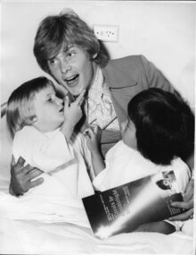 John Farnham holding a 1970 Carols program and two children who touch his face and jacket