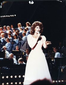 Woman with tall hairstyle and long pink gown sings to audience, with blue choristers behind her