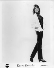 Smiling woman wearing light coloured jacket, plaid blouse, jeans and court shoes.