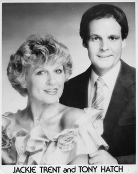 Seated woman and standing man wear odd smiles as they look to camera
