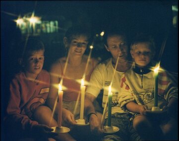 Family holding candles as they sit on the ground at Carols