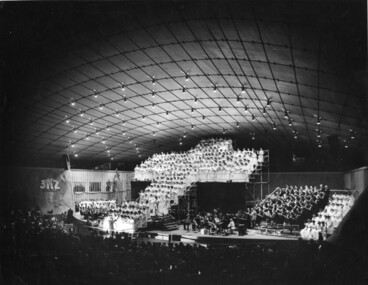 Choristers stand on raised platform in the shape of a Calvary cross with the orchestra and other choristers around them on stage