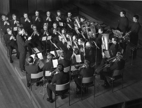 30 men playing musical instruments watch their conductor in a hall