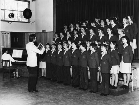 In a hall, a male conducts a choir of children whilst a woman plays piano.