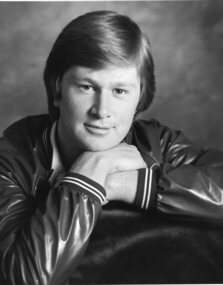 Denis Walter in pleather bomber style jacket rests his arms on the back of a chair