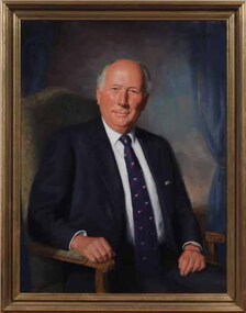 Man in blue suit, white shirt and patterned blue tie sits on a chair, turning to face towards the viewer
