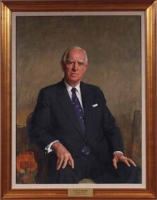 White haired man wearing a blue suit, white shirt and striped blue tie sits in a chair facing towards the viewer