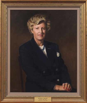 Fair haired woman in blue jacket with white top and blue beads, hands crossed in her lap, sitting on a chair looking towards the viewer