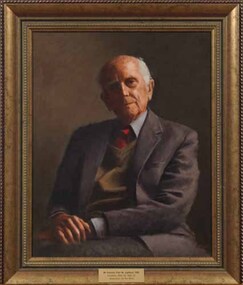 Elderly man in grey suit with green vest and red tie, with hands together on his lap to one side and tilted head looking outwards