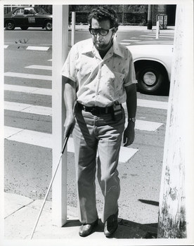 Bearded man uses white cane and sonic guide to walk over crossing